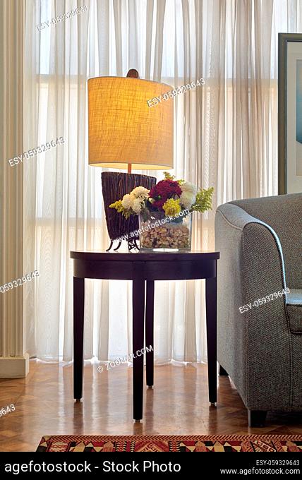 Interior shot of illuminated table lamp and colorful flowers planter on small dark brown wooden table on background of big window with white sheer curtains and...