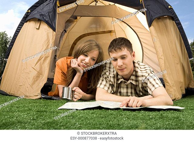 Photo of resting couple lying on green grass in tent and reading guide