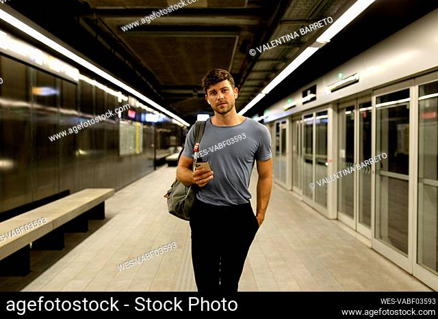 Handsome young man holding smart phone while walking with hand in pocket at illuminated subway platform