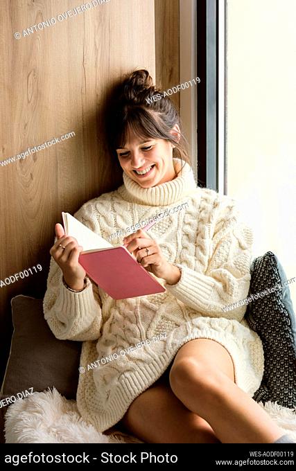 Smiling woman writing in book while sitting by window at home