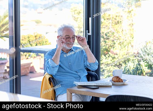 Senior man removing adjusting eyeglasses while sitting by table at home