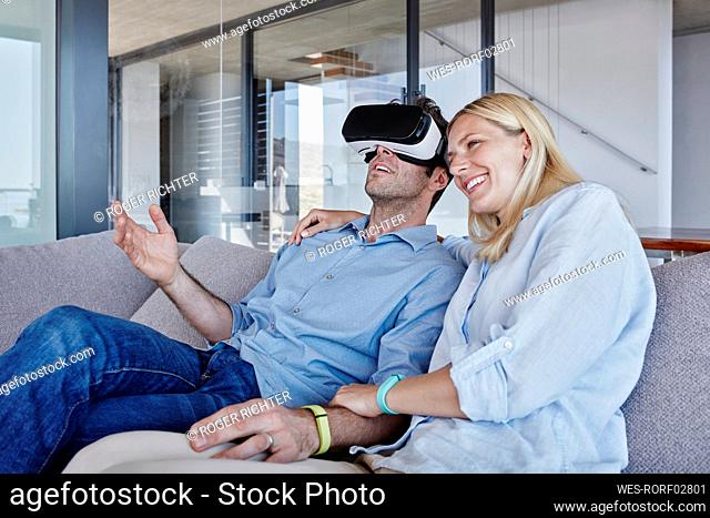 Smiling woman sitting by man wearing virtual reality simulator in living room