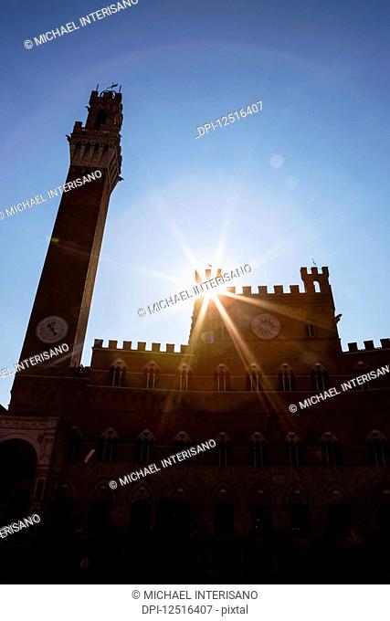 Backlit Palazzo Pubblico (town hall) and Mangia Tower with sunburst and blue sky; Siena, Tuscany, Italy