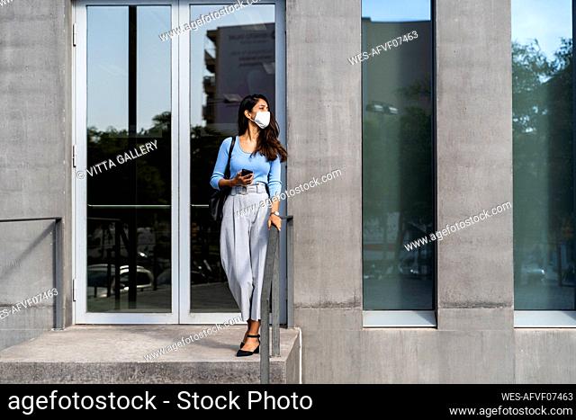 Young woman wearing protective face mask standing at entrance of building during COVID-19