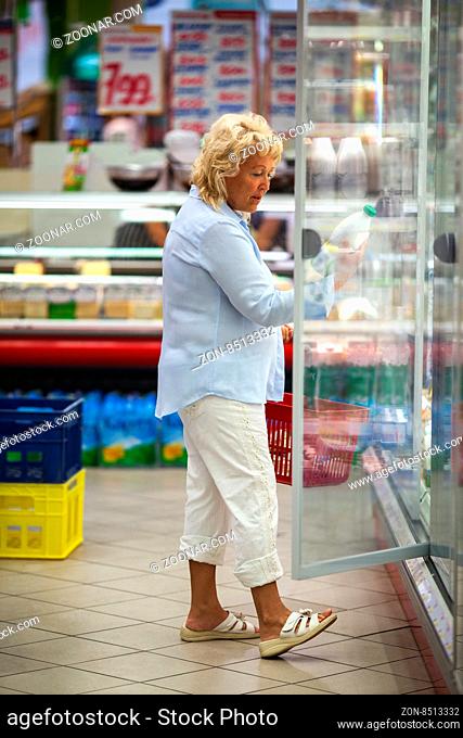 Senior woman choosing dairy in the store. She taking the bottle from the fridge and reading the label