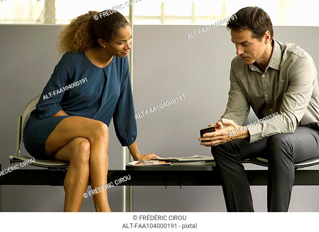 Professionals chatting in waiting room, man showing woman smartphone