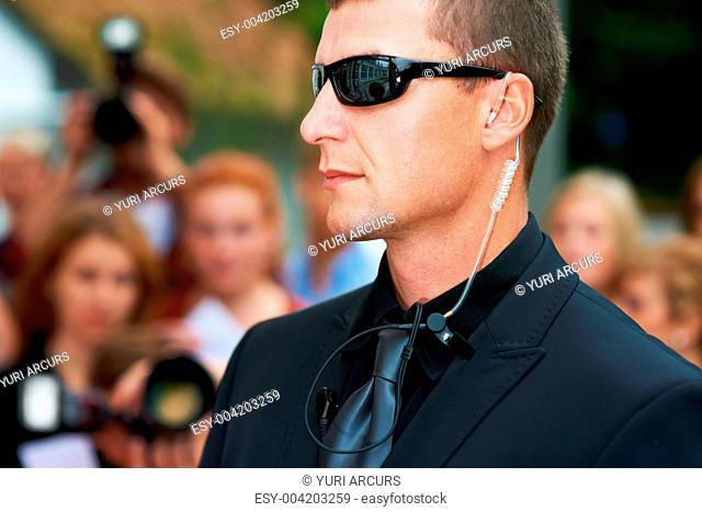 A security guard wearing dark glasses and an earpiece standing in front of the paparazzi
