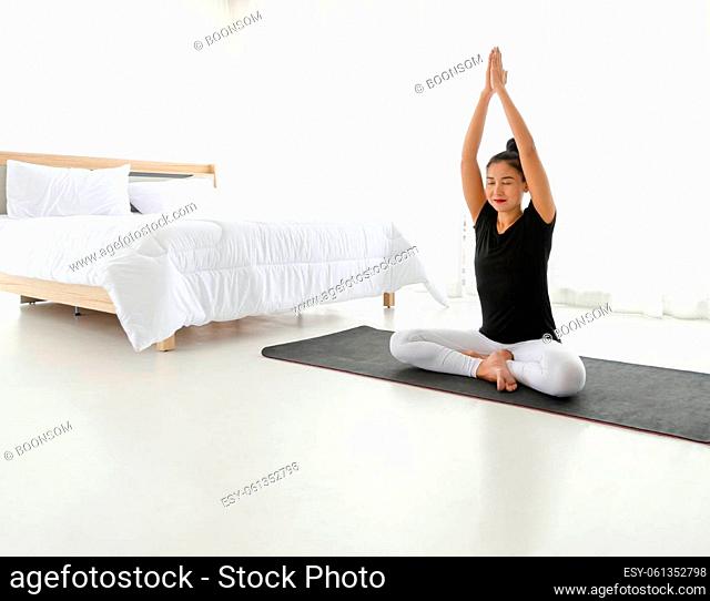 Asian women doing yoga meditating exercise at home, sitting in Easy seat pose or Sukhasana with raised hands above head pose in white bedroom
