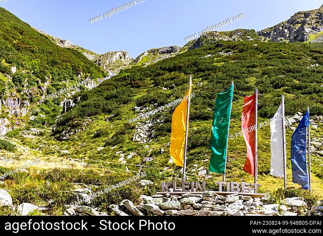 24 August 2023, Austria, Brandberg: Flags in the colors of Tibetan prayer flags stand on a hiking trail at the Zillergrund reservoir above the wooden...