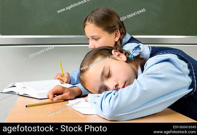Two girls sitting at desk. On is sleeping, other is writing something and reading a book. Side view