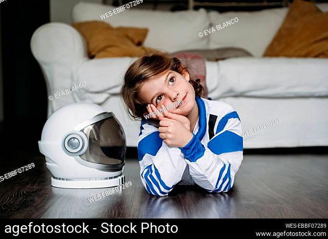 Thoughtful smiling girl wearing space costume lying by helmet on floor at home