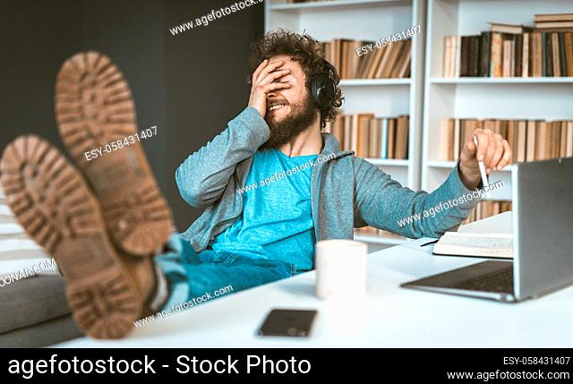 Facepalm. A young programmer works remotely at home. A young man smiles, covering his face with his hands sitting at a table in the office