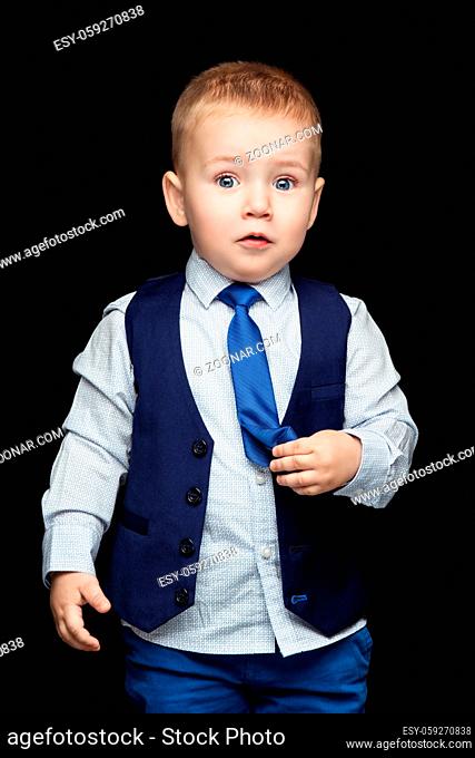 Cute little boy in blue business suit with surprised expression on face, touching his tie. Over dark background