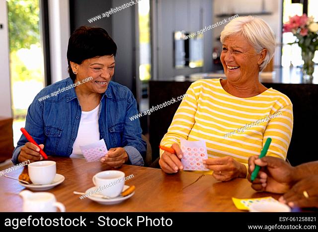 Cheerful biracial senior female friends with coffee and cookies on dining table playing bingo