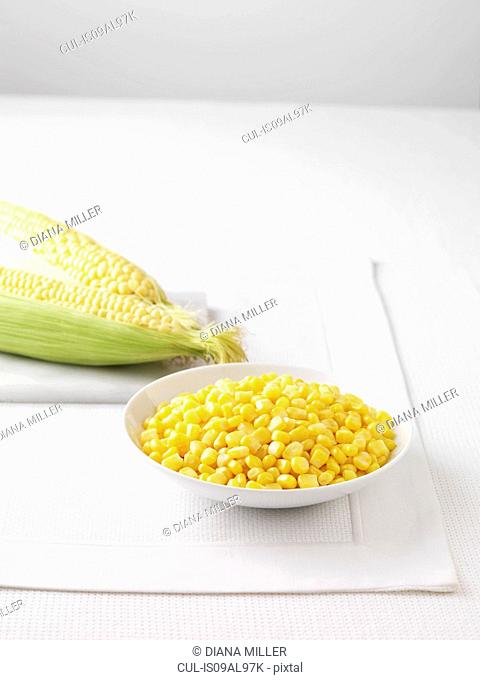 Raw corncobs on marble cutting board and bowl of boiled sweetcorn