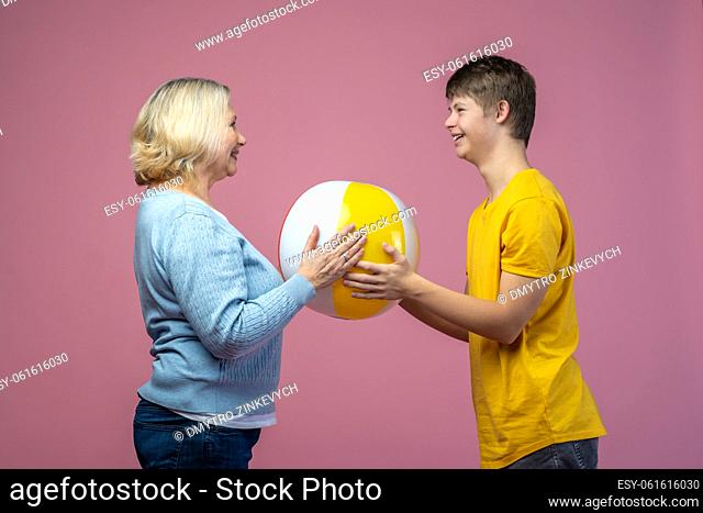 Communication. Joyful guy with down syndrome and middle aged woman standing sideways to camera opposite each other holding ball together