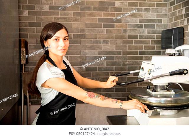 Beautiful chef woman wearing black apron using dough press machine to make flat pizza bread in kitchen at pizza store while looking at camera