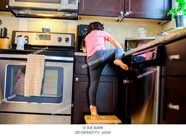 Young girl climbing onto kitchen work surface from stool, rear view