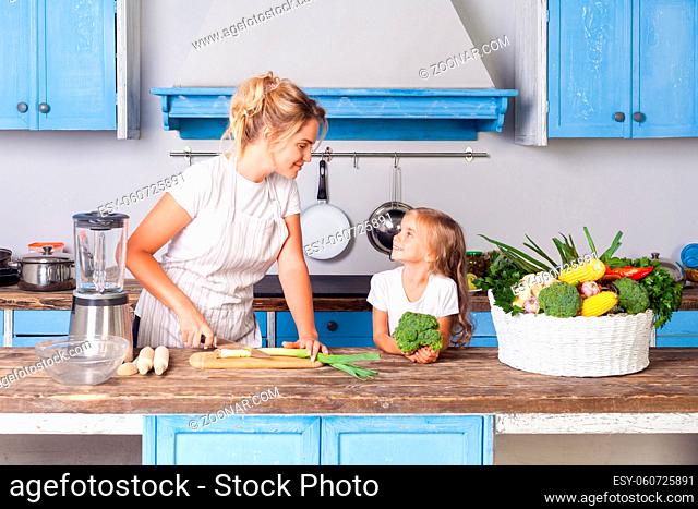 Happy pretty mother in apron chopping cutting green vegetable with knife and smiling to little daughter, woman cooking salad in kitchen
