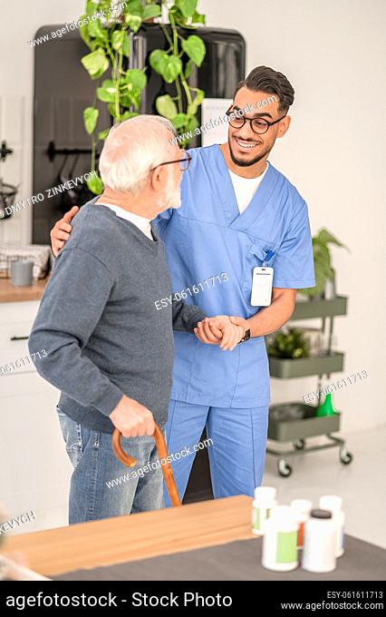 Smiling amiable young in-home nurse assisting his male patient to lean on the walking stick