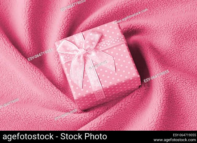 A small gift box in pink with a small bow lies on a blanket of soft and furry light pink fleece fabric with a lot of relief folds