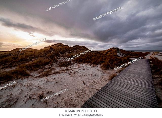 Deserted wooden boardwalk leading away through coastal dunes vegetation towards a glowing cloudy sunset sky in a moody evening landscape on Amrum