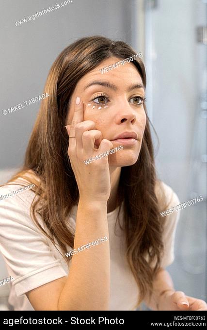 Young woman applying cream under eyes while standing at home