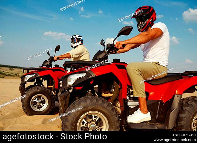 Two men in helmets and glasses ride on atv in desert. Male persons on quad bikes, sandy race, dune safari in hot sunny day, 4x4 extreme adventure, quad-biking