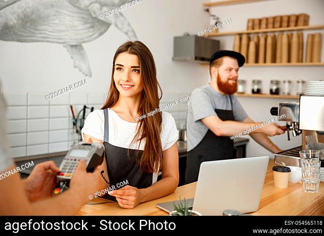 Coffee Business Concept - Beautiful female barista giving payment service for customer with credit card and smiling while working at the bar counter in modern...