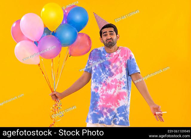 Portrait of sad young man standing with balloons and party horn in hands