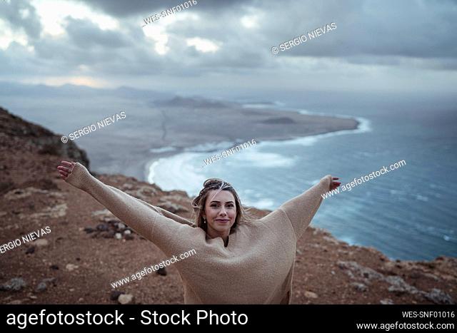Smiling woman standing with arms outstretched on mountain against Famara Beach, Lanzarote, Spain