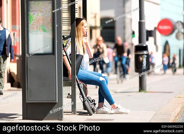 Casual caucasian teenager commuter with modern foldable urban electric scooter sitting on a bus stop bench waiting for metro city bus