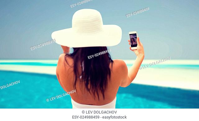 lifestyle, leisure, summer, technology and people concept - smiling young woman or teenage girl in sun hat taking selfie with smartphone over beach and swimming...