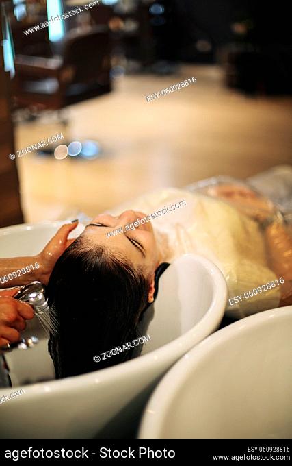 Cheerful young woman enjoying head massage while getting her hair washed by a professional hairdresser. Beauty care, hairstyling, fashion