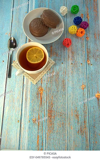 A cup of tea with a slice of lemon, a spoon and a plate of chocolate cake lie on a wooden table with a decoration of colorful balls. Close-up
