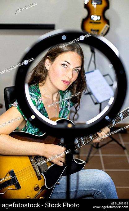 Confident woman with webcam light playing guitar while sitting at home