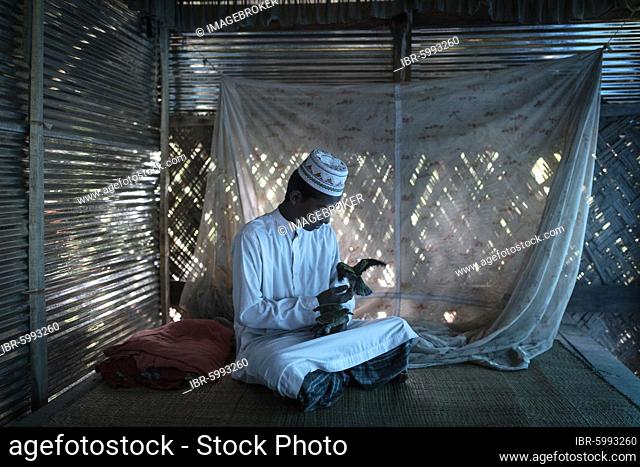 Ahmed on a raffia rug on the floor of a hut playing with two birds, wearing the white clothes of an imam, Mongla, Sundarbans, Bangladesh, Asia