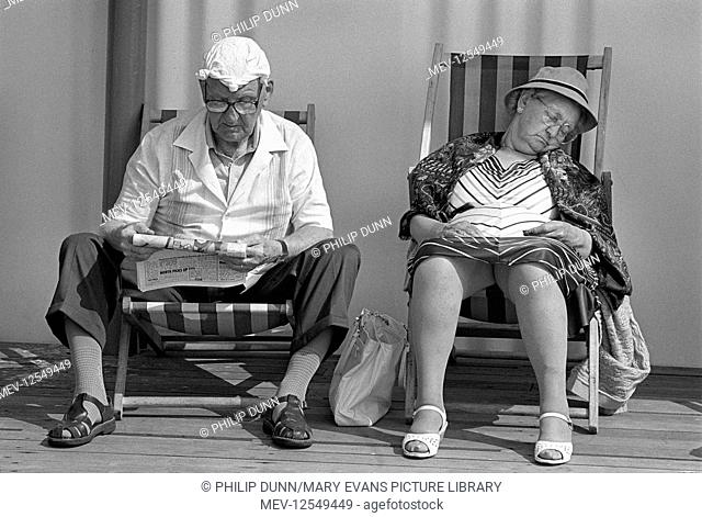 An elderly couple sit in deck chairs in a promenade shelter at Great Yarmouth, Norfolk, England. He reads the newspaper and wears a knotted handkerchief on his...