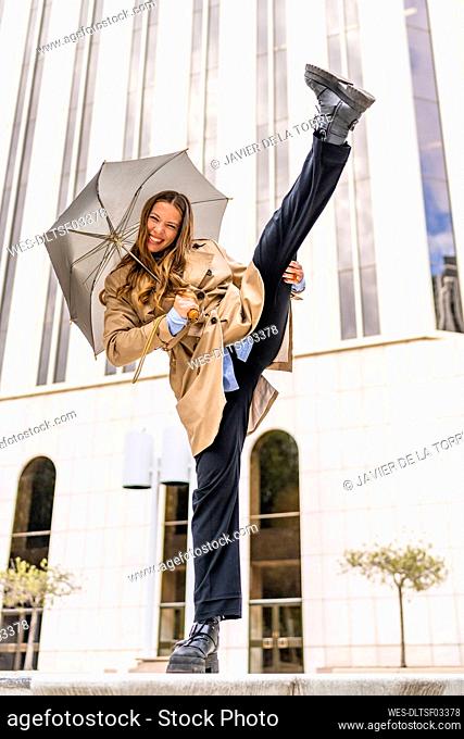 Playful businesswoman holding umbrella stretching leg in front of office building