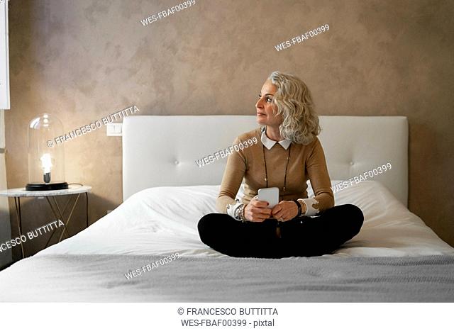 Mature businesswoman sitting on bed with mobile phone looking out of window