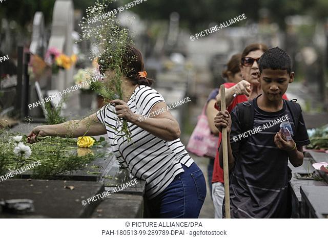 13 May 2018, Brazil, Rio de Janeiro: A boy with bank notes walks past a women laying down flowers on a grave at the Sao Francisco Xavier Cemetary