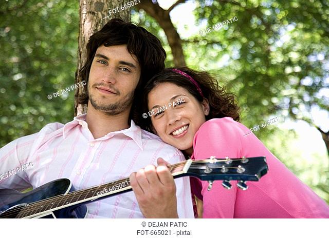 A young couple sitting underneath a tree and playing a guitar