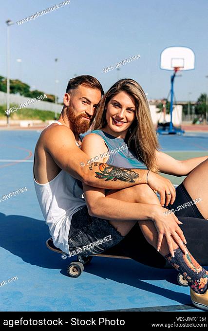 Smiling young woman sitting with boyfriend on skateboard at basketball court