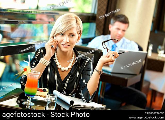 Young businesswoman sitting at table in cafe, talking on mobile phone, reading magazine