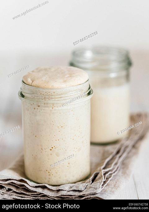 Two glass jars with wheat sourdough starters in different hydration levels. Starter 100 percent hydration in the foreground and starter with higher hydration in...