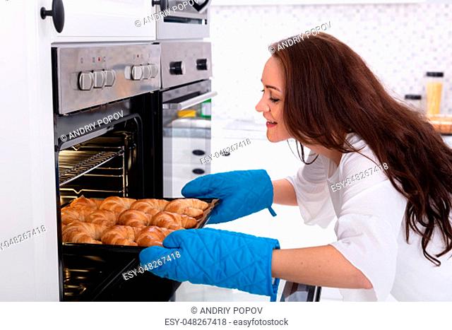 Side View Of A Happy Young Woman Removing Tray With Baked Croissants From Oven