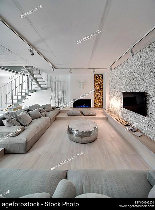 Contemporary interior with white walls. There is white stair with a metal railing, sofas with pillows, sparkle pouf, burning candlesticks and fireplace, TV