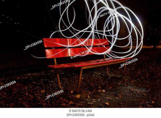 A red park bench in autumn with swirls of white light in the sky around it; Locarno, Ticino, Switzerland
