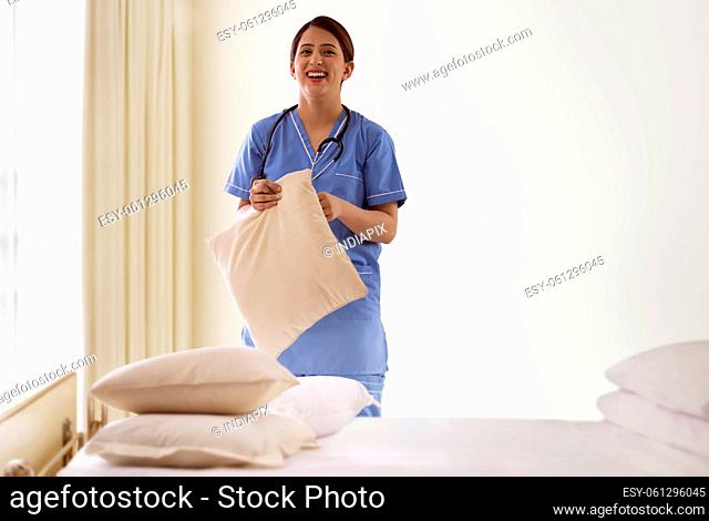 Portrait of a young nurse standing beside patient bed in hospital ward with pillow in hand