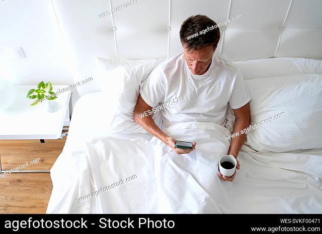 Mature man with smart phone and black coffee sitting on bed